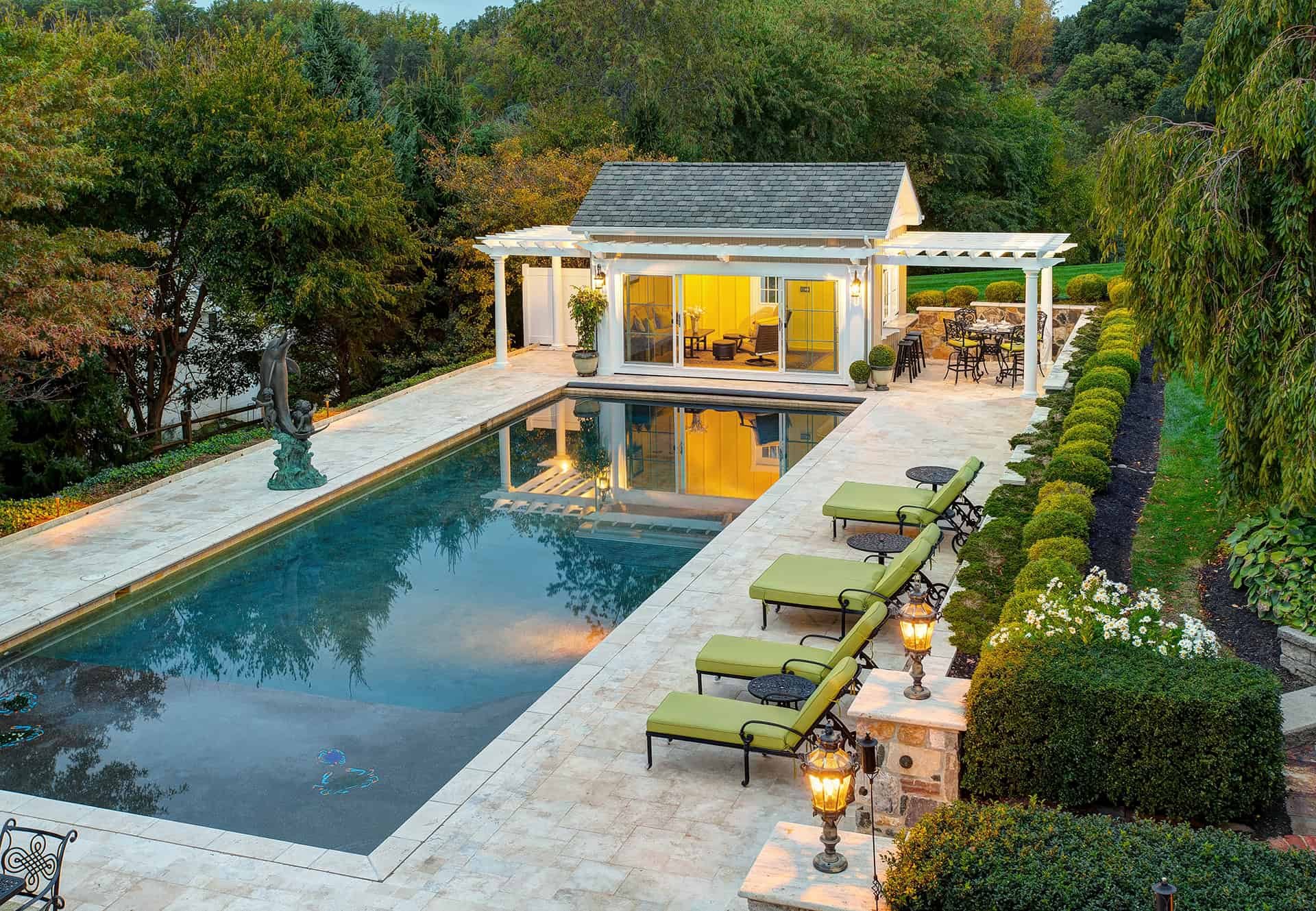 A luxurious backyard features a custom outdoor swimming pool, green cushioned lounge chairs, sculpted hedges, and a well-lit pool house with an outdoor seating area surrounded by trees and gardens.