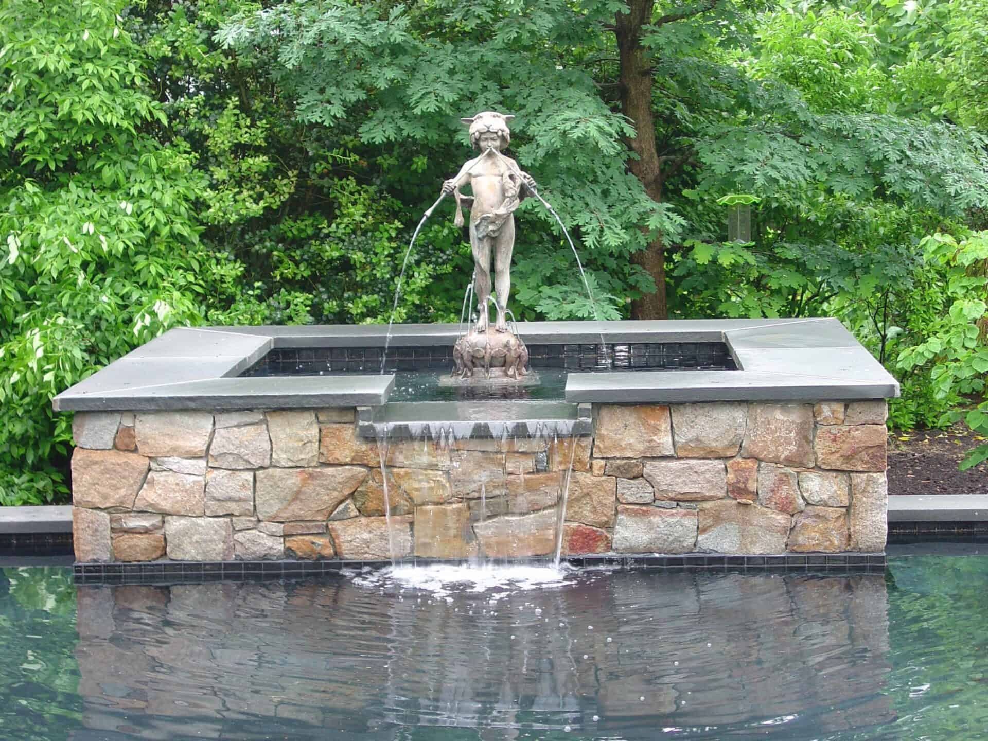 A stone fountain featuring a bronze statue of a child, holding two streams of water, stands among lush green trees and shrubs, with water cascading into a pool below—an elegant complement to custom outdoor swimming pools.